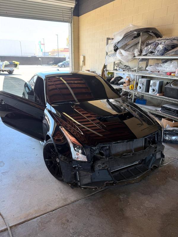 Black Cadillac torn down for collision repair just inside the roll-up door, its front bumper cover, grille and one headlight off and the radiator support exposed, with wrapped replacement panels stacked on the rack behind it