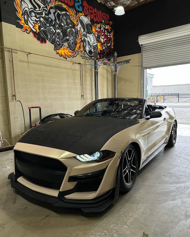 Ford Mustang convertible in champagne with a textured black vinyl hood, the matte hood panel stopping in a clean line at the fender and cowl edges against the gloss body; black splitter and wheels, top down, inside the shop