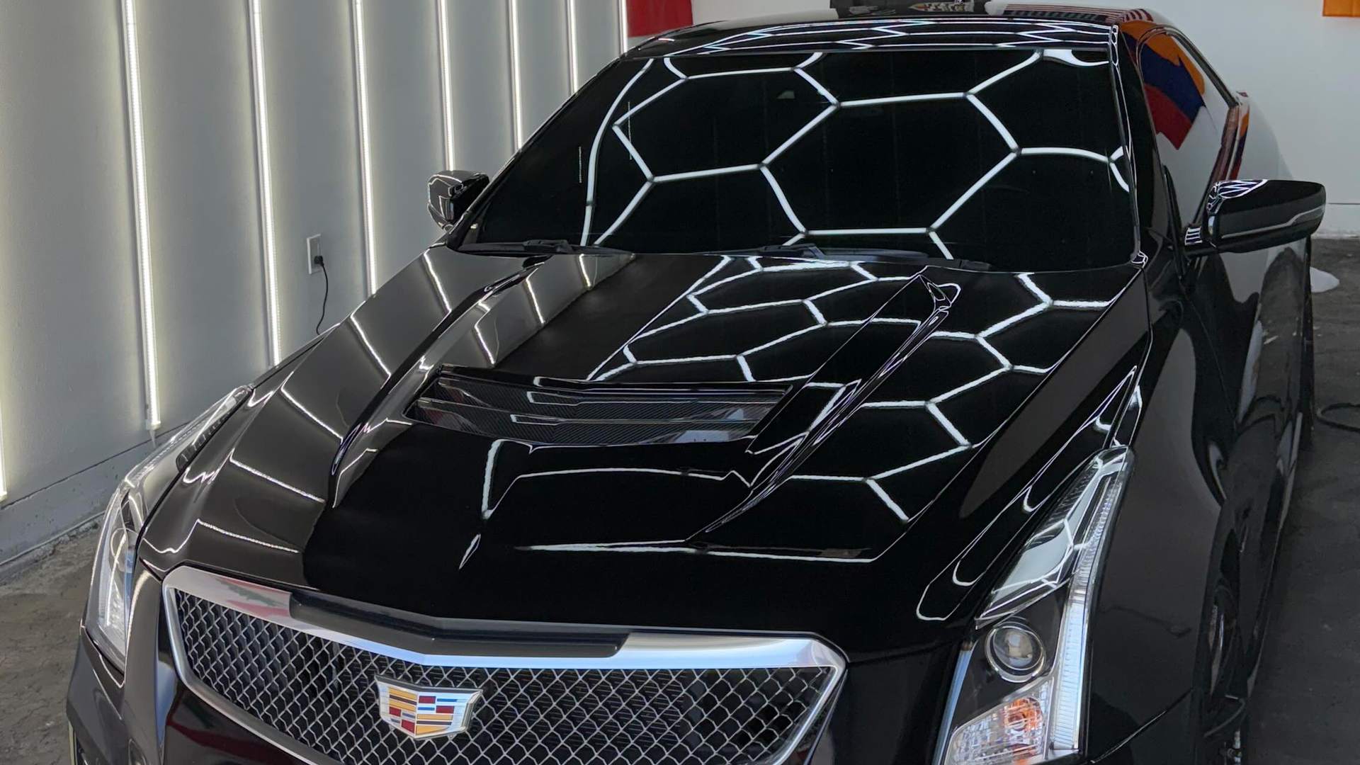 Cadillac ATS-V with its windows freshly tinted, shot head-on under the hexagon LED array so the array reflects whole across the gloss black hood and the darkened glass reads evenly dark corner to corner