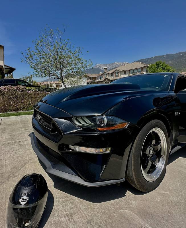 Close-up of the front corner of a gloss black Ford Mustang GT, the hood and fender carrying the sky and the ridgeline behind in one continuous reflection straight across the panel gap