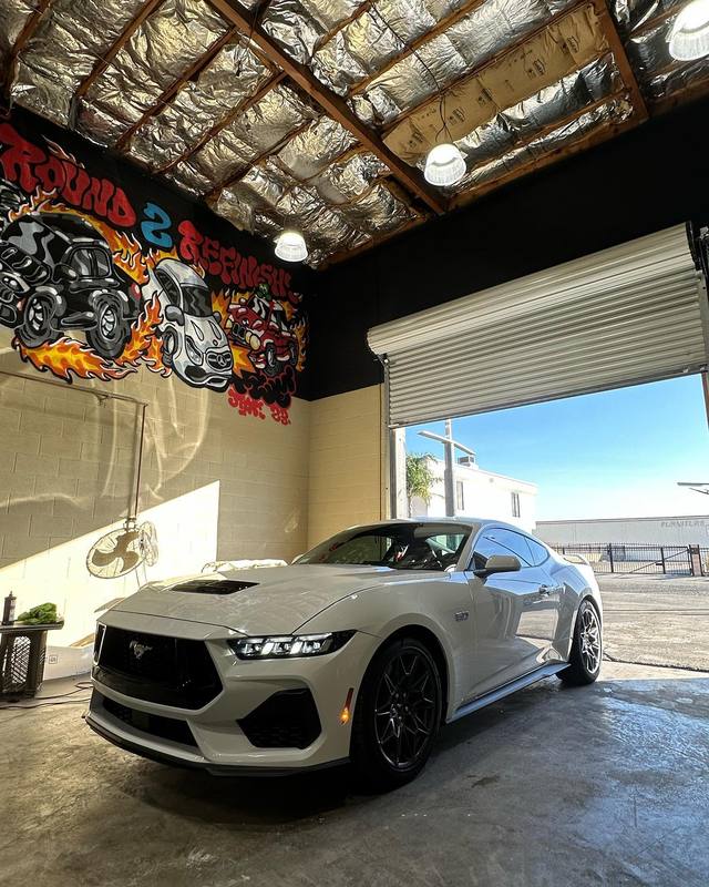 Ford Mustang GT in light grey with a black hood vent, black wheels and dark glass, parked inside the shop with the open roll-up door reflected the whole length of its flank