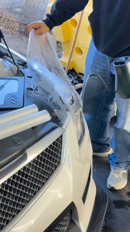 Paint protection film going onto the front corner of a white Cadillac ATS-V, the clear sheet floated on slip solution and drawn across the headlight and bumper before it is squeegeed down