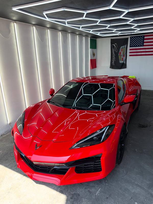 Chevrolet Corvette C8 Z06 in gloss red under the hexagon LED array in the restyling bay, the array mirrored whole across the hood, fenders and windshield with the hexagons holding their shape over every curve