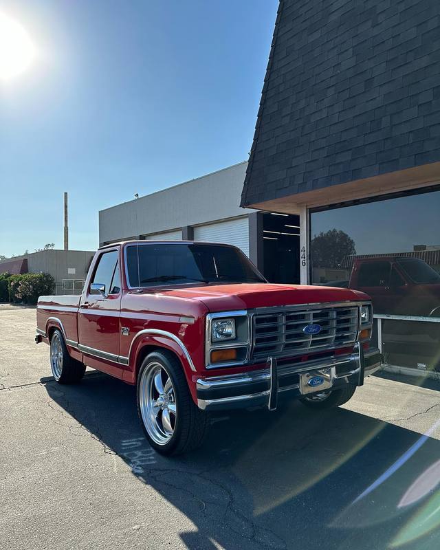 Red 1980s Ford F-150 parked outside the shop, red over a white lower body with bright chrome on the grille, bumpers and side moldings and the red holding a hard sun reflection along the bed side