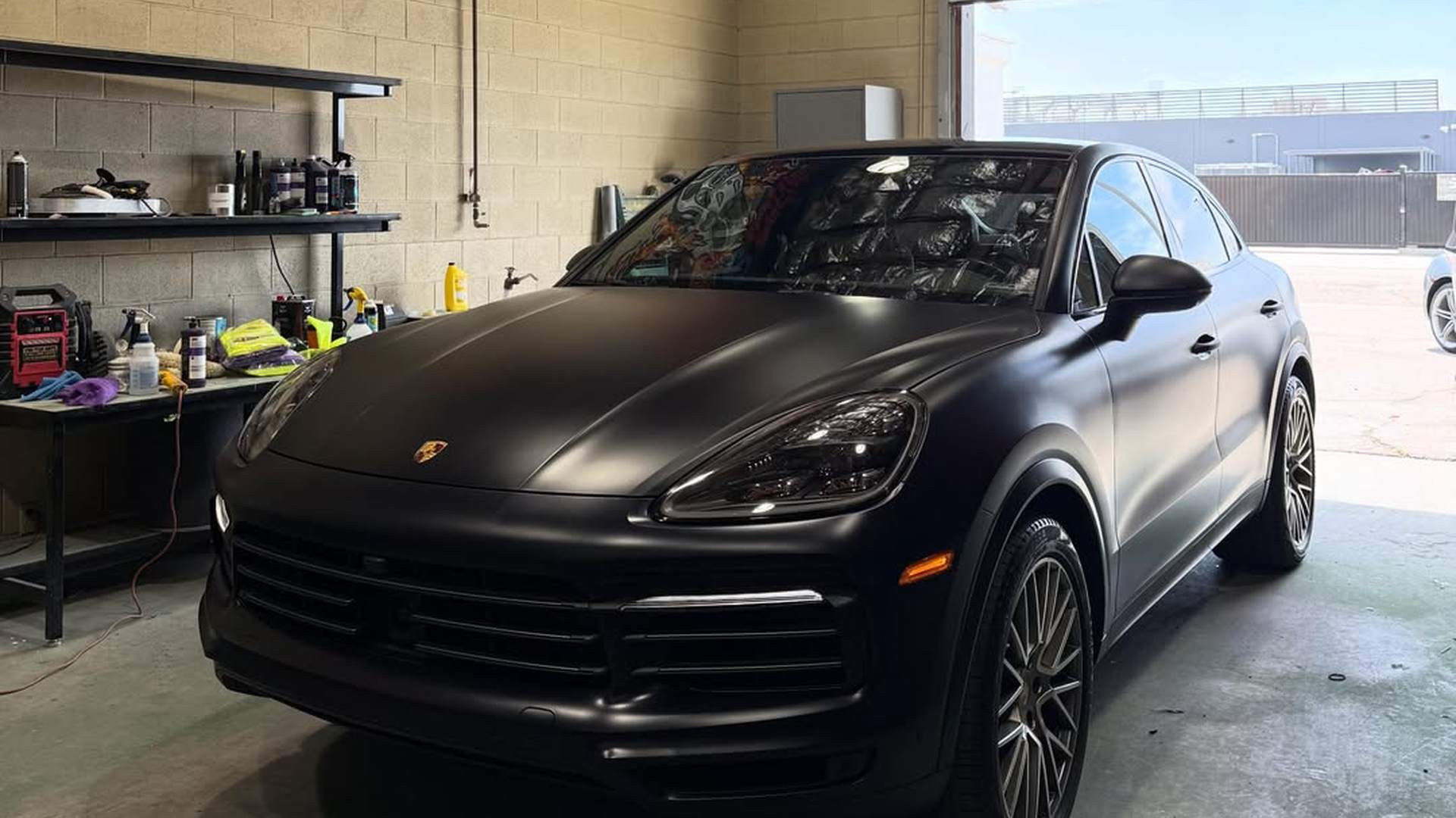 Porsche Cayenne Coupe after a Peel Clear color change, the satin black running unbroken over the hood, bumper, fenders and doors and around the headlights with no seams or cut lines anywhere; parked under the flame mural in the shop