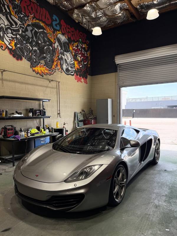McLaren MP4-12C in silver parked inside the shop beside the workbench, the low nose and the door intake picking up the daylight from the open roll-up door