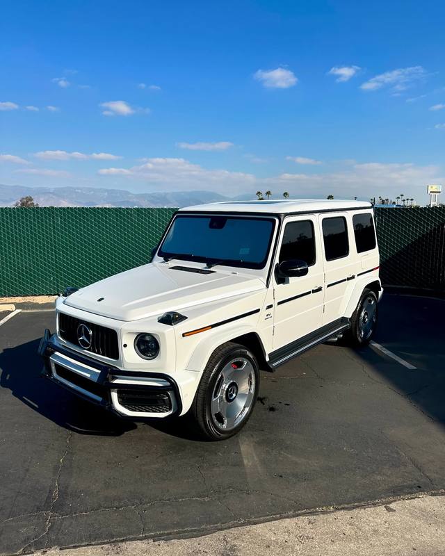 Mercedes-AMG G63 in gloss white with dark glass and black window frames and side moldings, the chrome grille, brush guard and wheels left bright against it, parked in the lot with the mountains behind