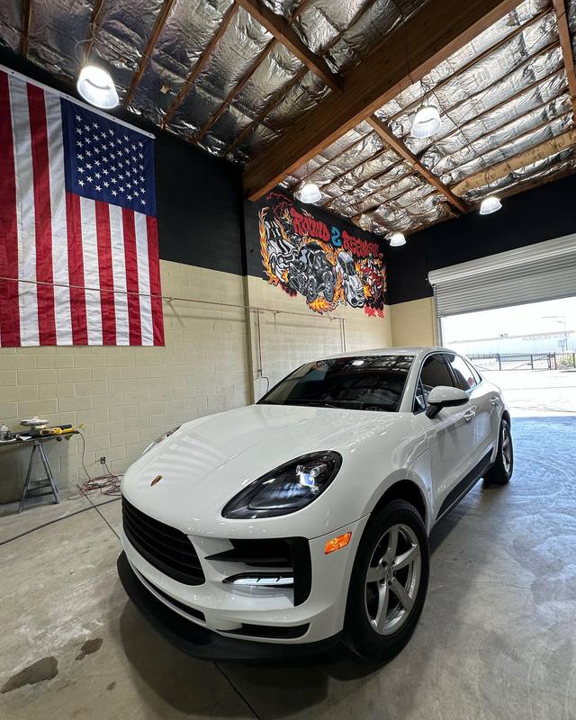 White Porsche Macan parked inside the shop under the American flag and the flame mural, the sealed floor throwing a soft reflection of the car back up beneath it