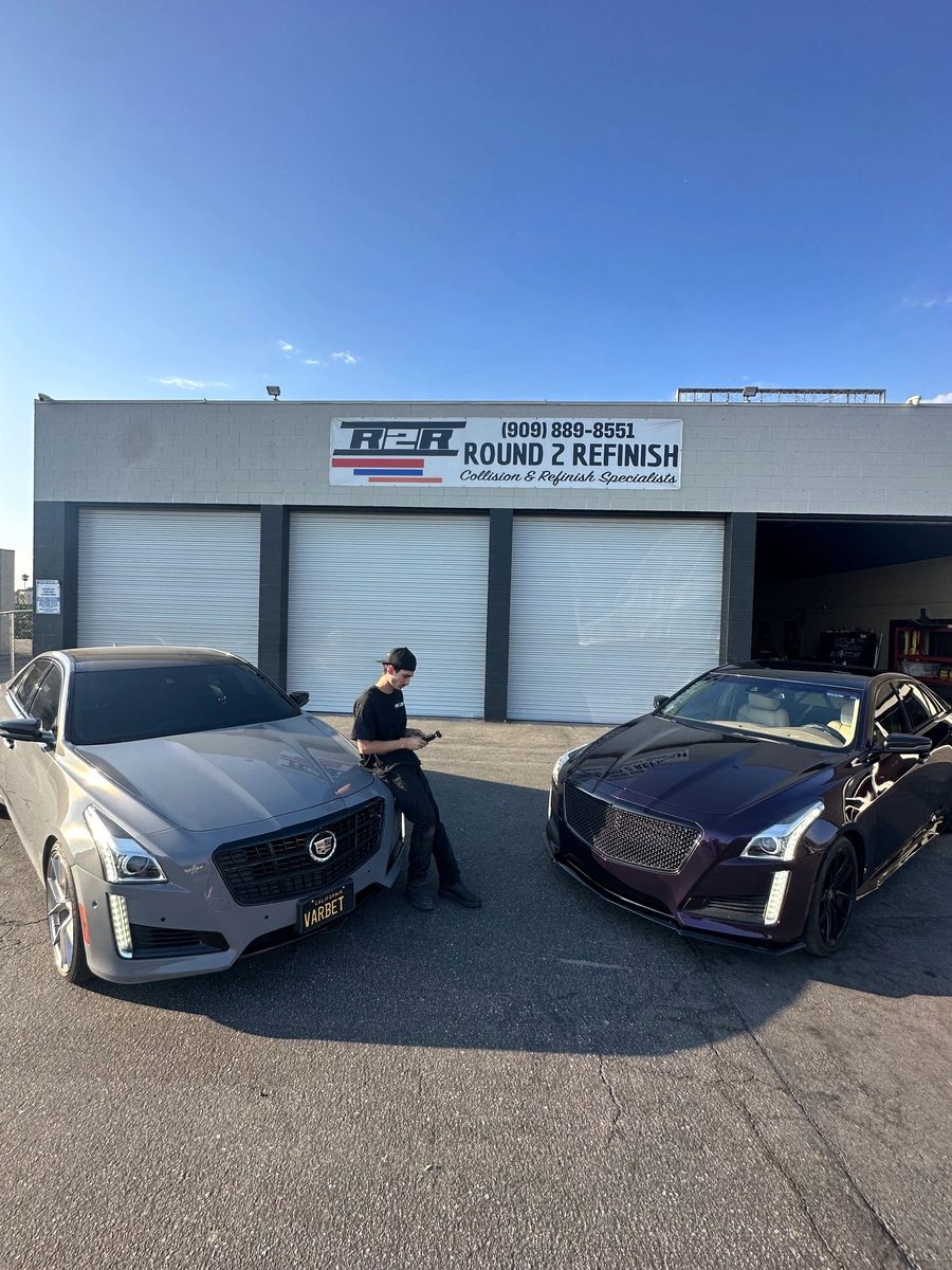 Vasken Hovannesian standing between two Cadillac CTS sedans outside the shop, a silver one on his right and one on his left whose hood, grille and fenders shift from deep purple to black as the light moves across them