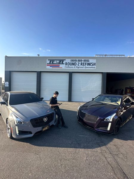 Vasken Hovannesian standing between two Cadillac CTS sedans outside the shop, a silver one on his right and one on his left whose hood, grille and fenders shift from deep purple to black as the light moves across them